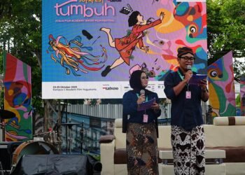 Two presenters stand on a stage with microphones, a colorful festival banner behind them reading 'tumpuh' and event details.