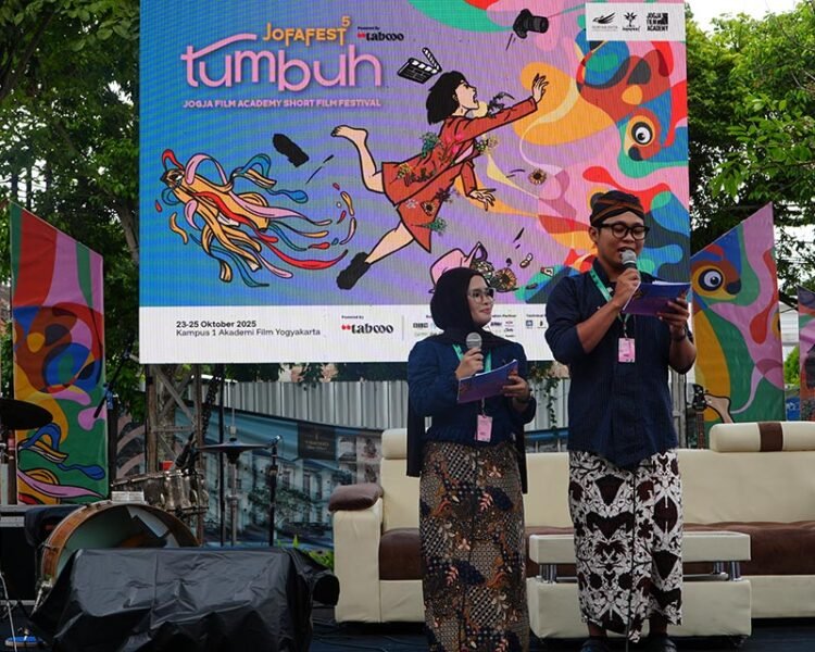 Two presenters stand on a stage with microphones, a colorful festival banner behind them reading 'tumpuh' and event details.