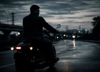 A motorcyclist on a dark motorcycle rides along a wet, reflective road at dusk with city lights in the distance.