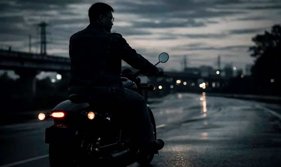 A motorcyclist on a dark motorcycle rides along a wet, reflective road at dusk with city lights in the distance.