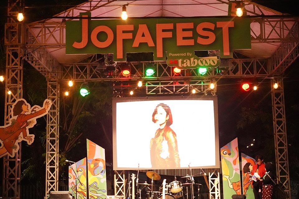 Jofafest 5 Stage at JOFAFEST with a large screen displaying a portrait, drum set, guitarist, and colorful festival decorations.