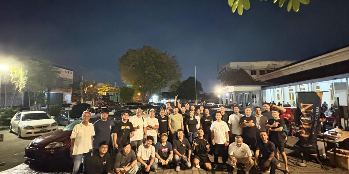 Large group of people posing for a group photo in a dimly lit parking lot at night, with cars parked along the street and a building on the right.