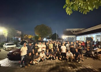 Large group of people posing for a group photo in a dimly lit parking lot at night, with cars parked along the street and a building on the right.