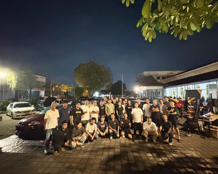 Large group of people posing for a group photo in a dimly lit parking lot at night, with cars parked along the street and a building on the right.