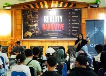 Presenter speaks into a microphone at a conference stage with a large 'From Reality to Narrative' banner behind him/her.
